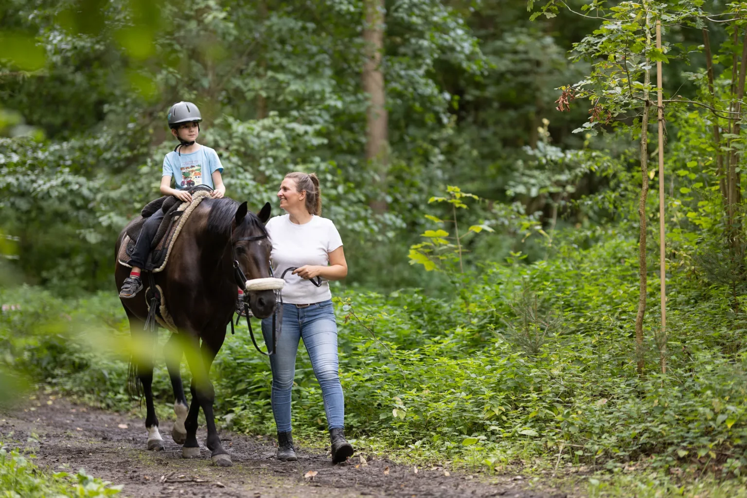 Ein Kind reitet auf einem Pferd durch den Wald, begleitet von der Therapeutin