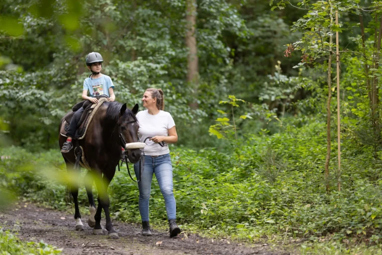Ein Kind reitet auf einem Pferd durch den Wald, begleitet von der Therapeutin